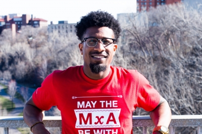 A man with his arms draped on a concrete wall in New York City wearing a red shirt that says MxA = Force tech humor for May the Force Be With You.