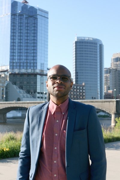 A young man stands in front of a bridge and cityscape under a blue sky in Grand Rapids, Michigan. He is wearing a suit.
