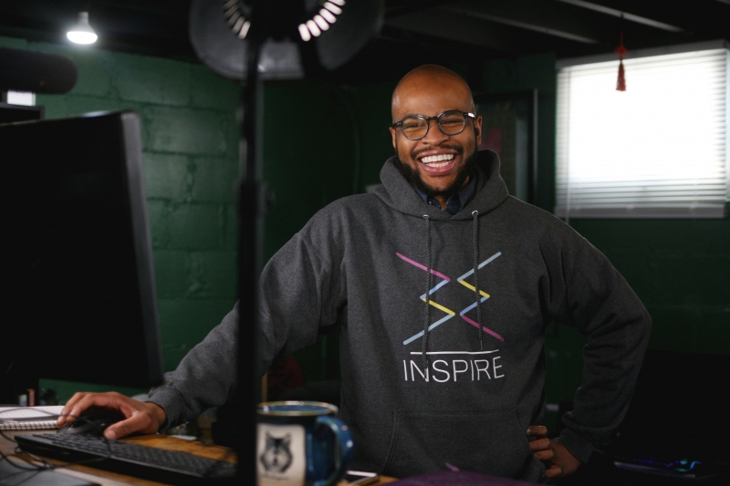 A young man wearing an Inspire sweatshirt smiles wearing glasses near a computer with a coffee cup