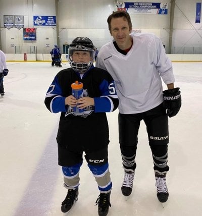A dad and his son on a Kansas ice rink in hockey gear with the Midwest Championship sign behind them and the son holding a water bottle. They both are suited up in their hockey gear.
