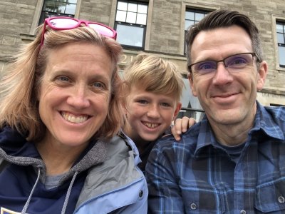 A mom, 10-year-old son, and dad outside a stone building with a blue sky. They are a smiling family wearing fall jackets..