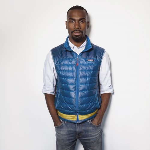 A man stands in a puffy vest with his hands in his pockets looking at the camera in front of a white background.