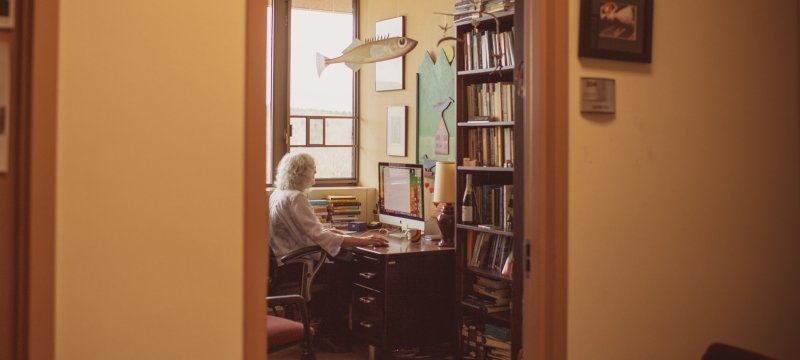 A woman sits at a desk inside an office working at her computer as the camera looks in outside from the hallway. There is a hanging wooden fish sculpture, books, and a window in the background at a University.