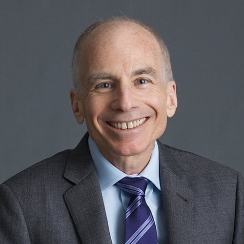 An older man wearing a suit and tie smiles at the camera with a blue background behind him. 