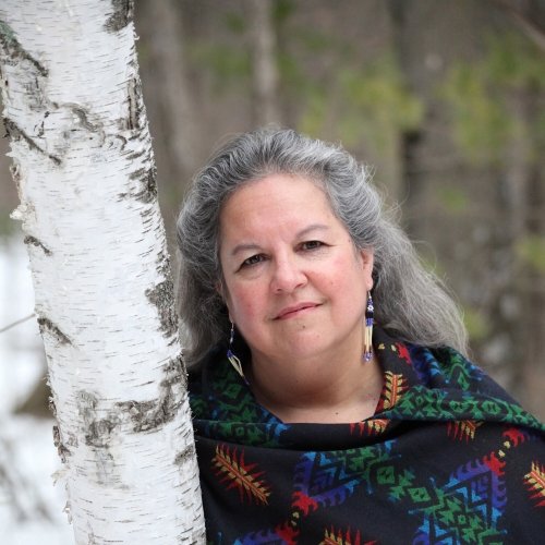 A woman leans against a white birch tree in the forest, hair blowing in the wind as she smiles at the camera.