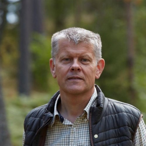A man in a puffy vest with gray hair looks at the camera with a forest in the background.