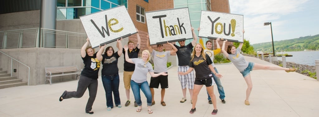 A group of students in unique poses holding three signs with signatures; the signs have the words "We Thank You" with one word per sign.