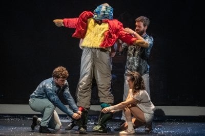 An actor stands on stage with arms outstretched while three others assist with putting on winter gear.