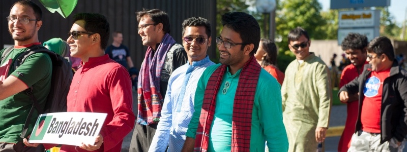 Bangladesh students walking in the Parade of Nations; some are wearing traditional dress and one student is holding a sign that has their country name and flag.