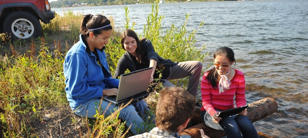 Faculty and students sit on the edge of the Keweenaw Waterway while looking at laptops discussing.