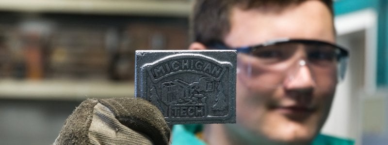 A man holds up a metal casting that reads "Michigan Tech."