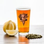 A pint glass full of beer sits beside a sliced lemon and a pile of pelletized hops.