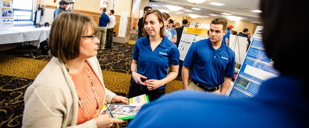 A young woman and man in blue shirts in front of a poster in a ballroom exhibit talk to a woman holding a Design Expo booklet and clipboard
