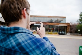 A young man in a blue plaid shirt records video on his phone, his back is to us, he is outside facing a building with windows.