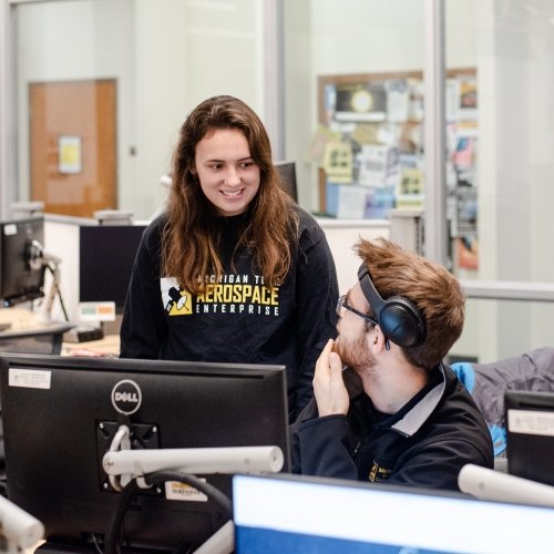 a female student with Michigan Tech Aerospace on her shirt talks to a male student sitting at a monitor wearing head phones like ear muffs