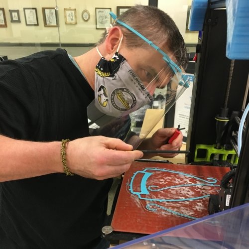 A man with a Michigan Tech Huskies face mask and a face shield on campus with a 3D printer printing PPE face shields.