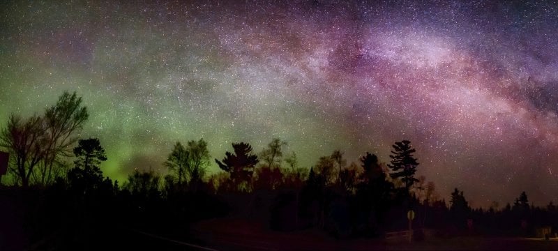 the milky way in the Keweenaw Peninsula with northern lights shimmerrs over a darkened forest treeliine.
