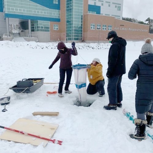Five people in winter clothes on the ice take a water sample, with one pulling a net out of a hole in the water near Michigan Tech's Great Lakes Research Center