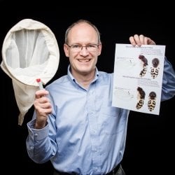 A man holds up a butterfly net and fruit fly closeup photo smiling with a black background.