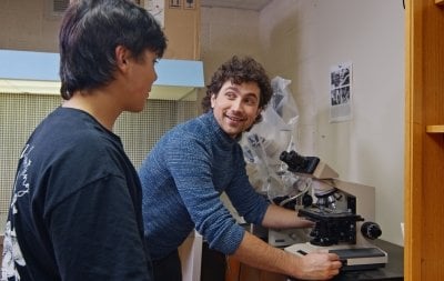 An undergraduate researcher stands at the microscope working with a first-year student in the College of Forest Resources and Environmental Science fungi lab at Michigan Tech.