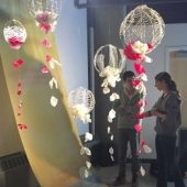 Two students standing inside the Rozsa Gallery discussing an exhibit of a long piece of fabric that is draped from the ceiling with a river and sunset painted on it. Above them is an installation with balloon-shaped pieces made with wire and simulated flower petals.