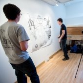 Two students work on a large drawing on a studio wall while a faculty observes and a camera records.