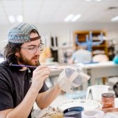 Student sits at a workbench in the ceramics studio painting a sculpture piece.