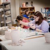 Three students sit at a workbench painting sculptures.