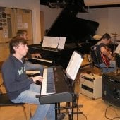 Student playing piano while other students play guitars in the Instrumental Rehearsal Room.