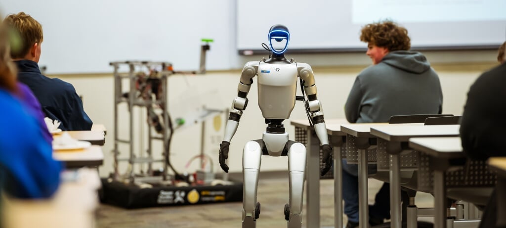 Students watching a humanoid robot walk between tables in a classroom