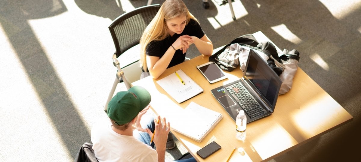 Two students studying together at a table in the sunlight in a library with a laptop and a tablet.