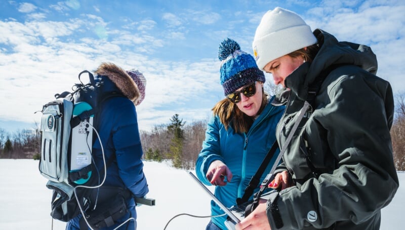 Two people review a tablet connected to the backup of a third person who is reviewing a different device. They are in a snowy field during a sunny day.