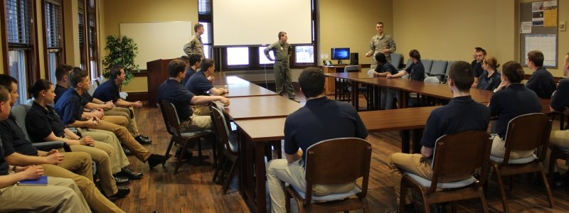 A large group of cadets sit inside the ROTC building while listening to peer present.