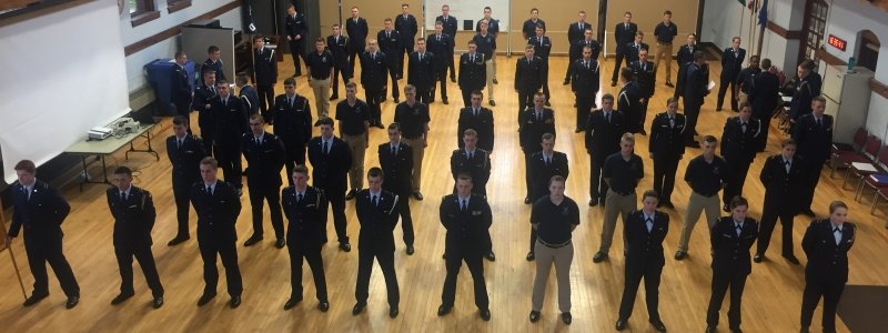 Seated cadets in uniform watch cadets in dress uniforms give a rifle demonstration