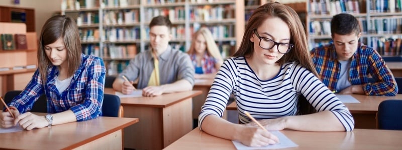 Students, two males and three females, sit in a library at desks taking a written exam.