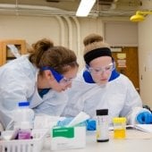 Five high school students in labcoat, goggles, and gloves working on an experiment in a lab.