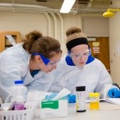 Five high school students in labcoat, goggles, and gloves working on an experiment in a lab.