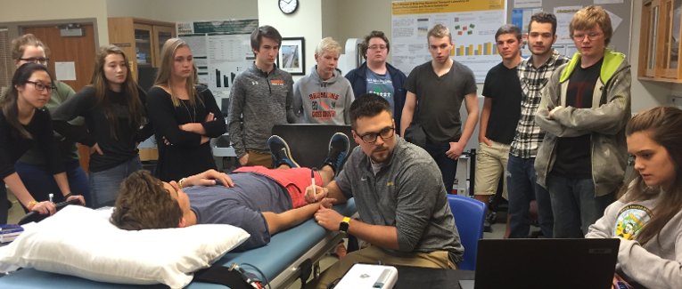 Students stand around a medical bed watching two researchers collect data.