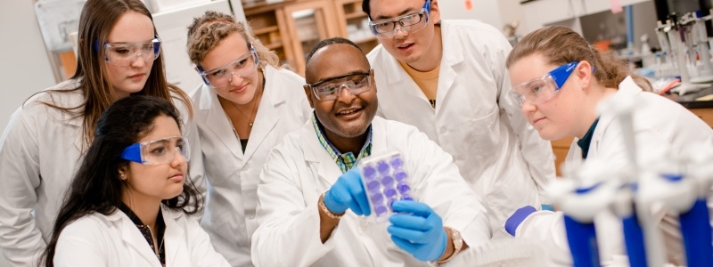 Dr. Tubman with students in researcher lab raising up a sample and pointing.