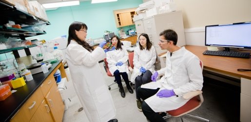 Faculty standing in front of three seated students in a lab with lab coats, gloves, and safety glasses.