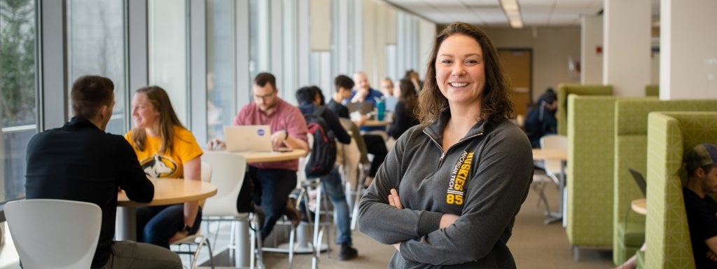 Smiling student standing with arms crossed in front of other students studying.