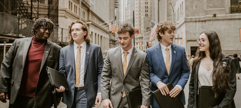 Five students in suits walking down a city street.