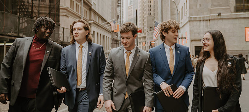 Five students in suits walking down a city street.