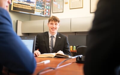 APMP student sitting at a desk with an open book, smiling at two people in the foreground.