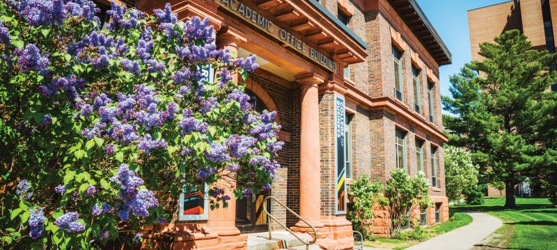Exterior of the Academic Office Building with a lilac bush blooming next to the entrance.