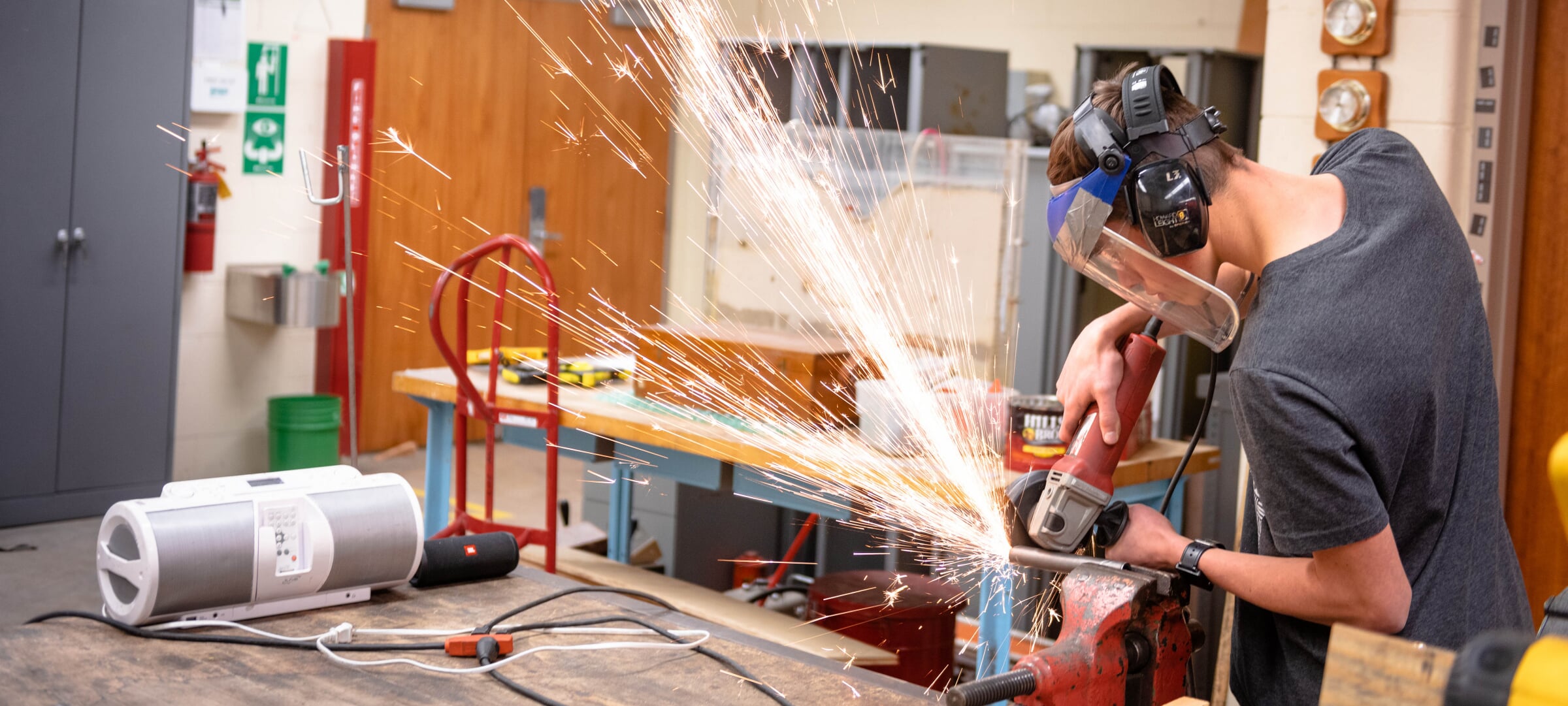 A student using a angle grinder on a pipe in a vice causing sparks to shoot out away from them.
