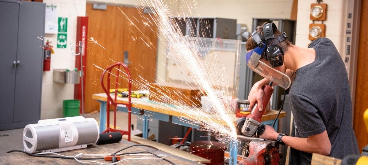 Graduate student in a lab with protective gear using a metal cutter to cut a pipe with sparks flying away