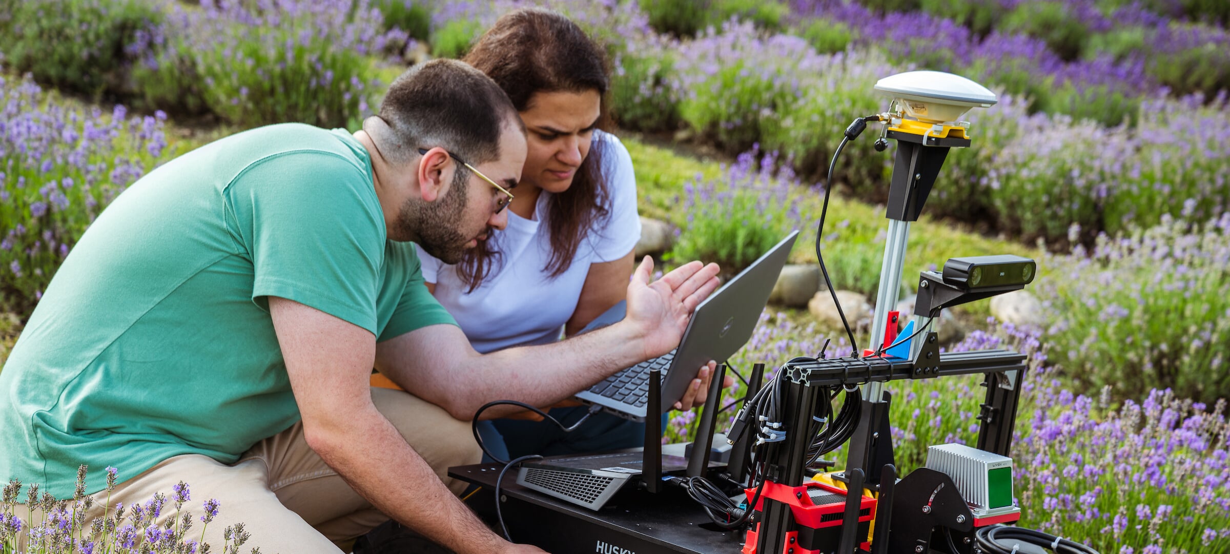 Two students in a field of lavender looking at a laptop while working on a robot
