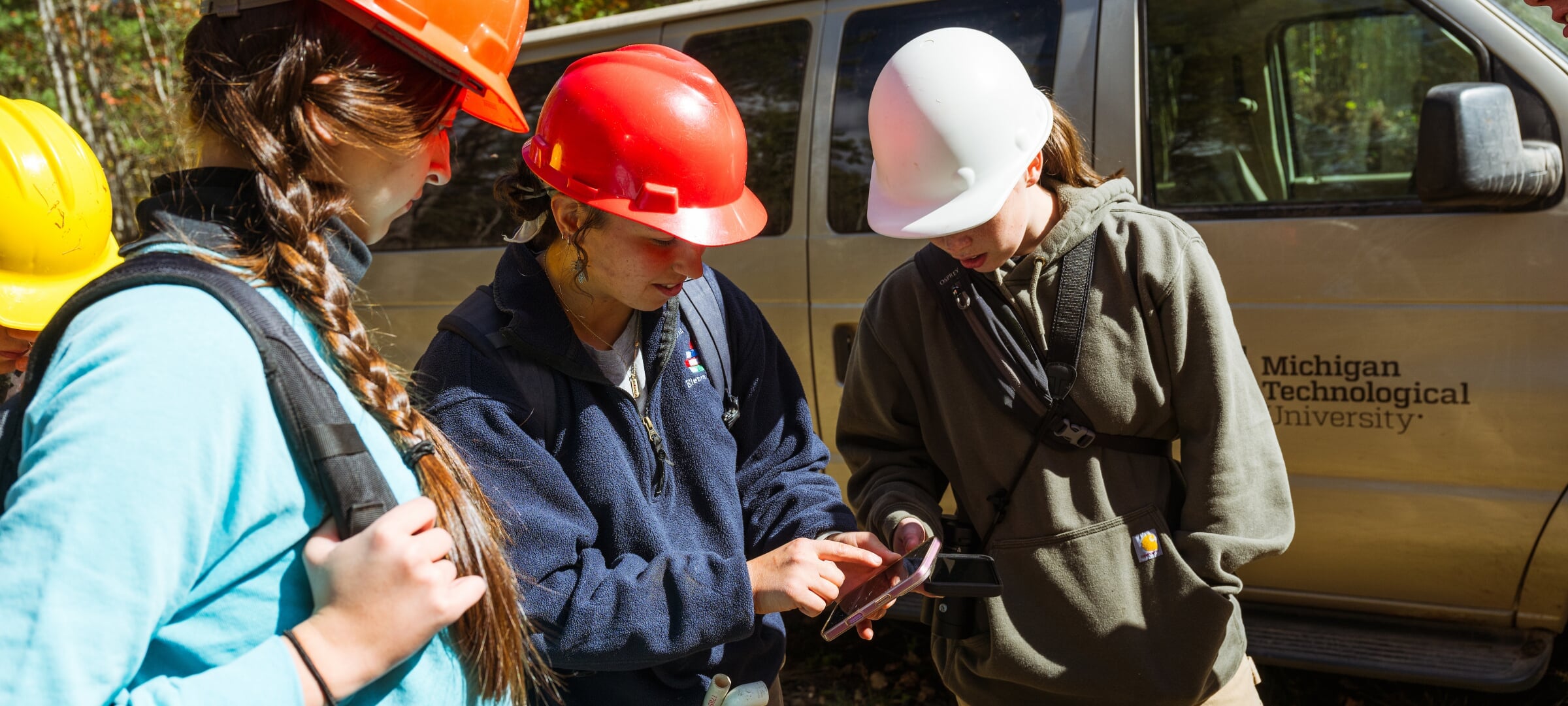Three Michigan Tech students in hard hats collaborating over a smartphone in the field.
