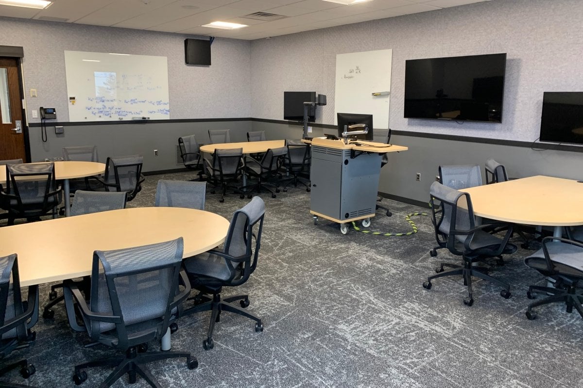 ChemSci 215 showing 4 group tables, each surrounded by 6 chairs with casters and a monitor mounted on the wall. Two white boards are on the wall and a larger monitor is mounted near the lecture station.